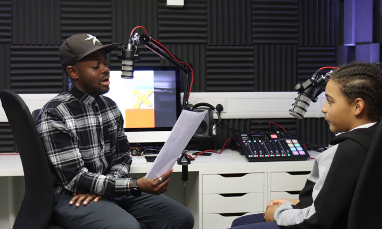 Young man holding a piece of paper and talking into a microphone, sat opposite a young person at a desk in a studio