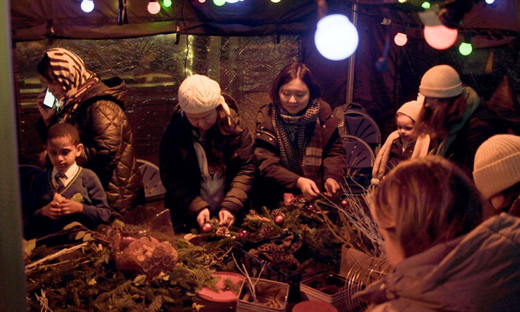 People gathered round a table with festoon lights above it making Christmas wreaths