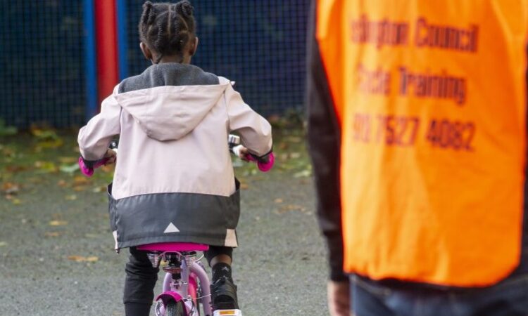 A young girl in a light pink jacket riding a bright pink bike with a man in high vis next to her