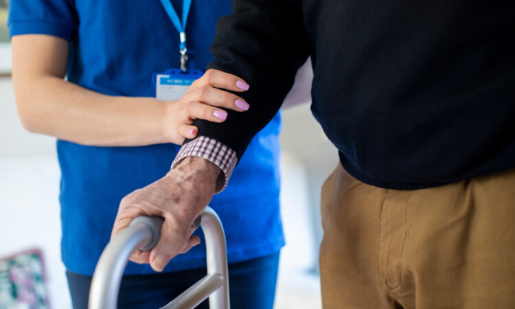 Close Up Of Senior Man With Hands On Walking Frame Being Helped By Care Worker