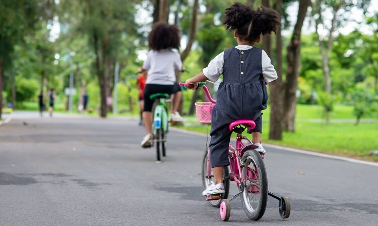 Photograph: two children cycle in a park; the younger girl has stabilisers on her bike.