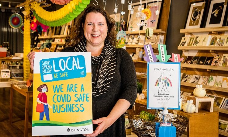 Photograph: Interior of The Pretty Shiny Shop, with owner Georgina standing in the foreground, holding a placard reading: "Shop Local" "We are a Covid Safe Business".