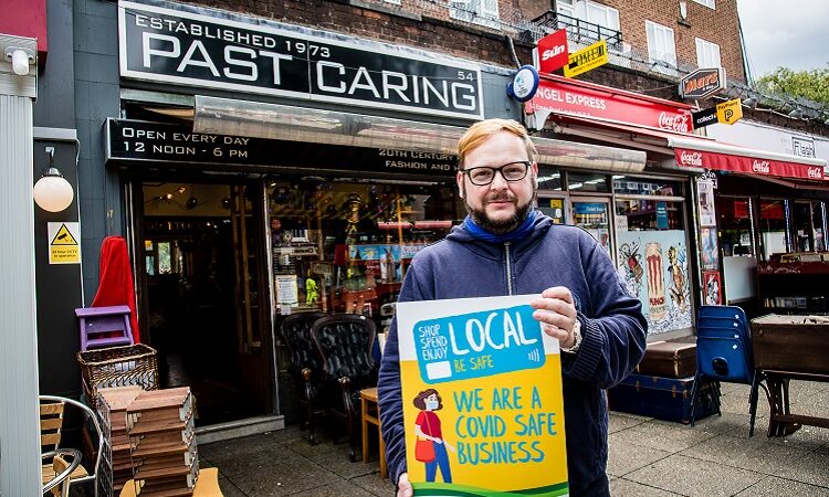 Photograph of the exterior of "Past Caring" shop, featuring staff member James. James is holding a placard reading: "Shop Local" "We are a Covid Safe Business"