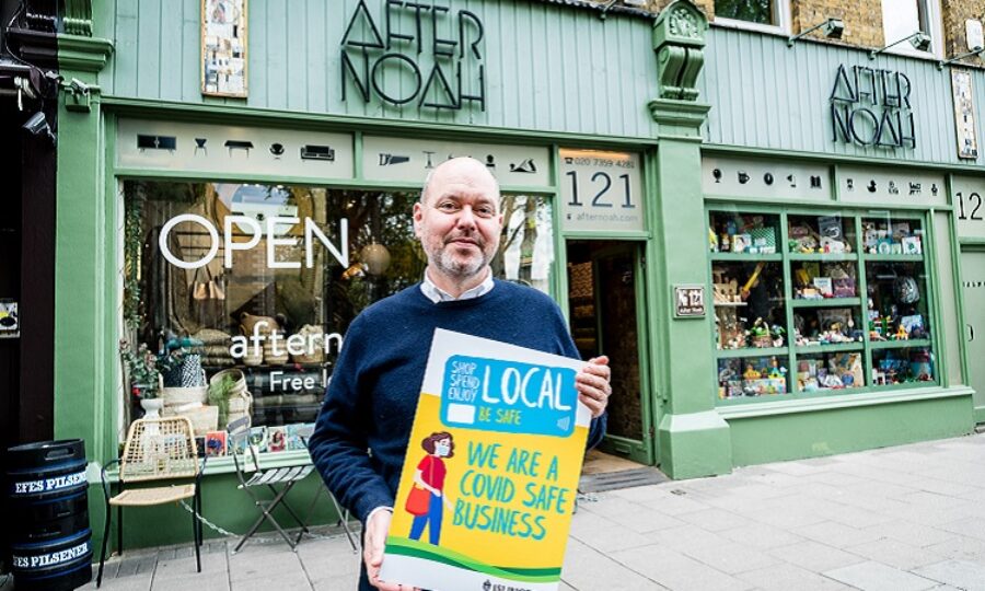 Photograph: exterior of "After Noah" shop. Part owner and manager Simon Tarr stands in front, holding a placard reading: "Shop Local" "We're a Covid Safe Business".