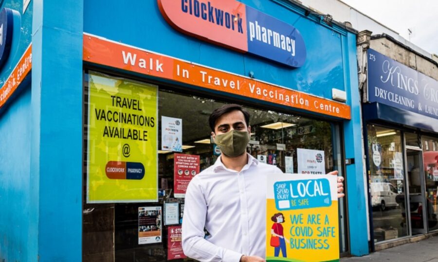 Photograph: exterior of Clockwork Pharmacy. Manager Vishal stands in front, wearing a face mask and carrying a placard reading: "Shop Local" "We are a Covid Safe Business".