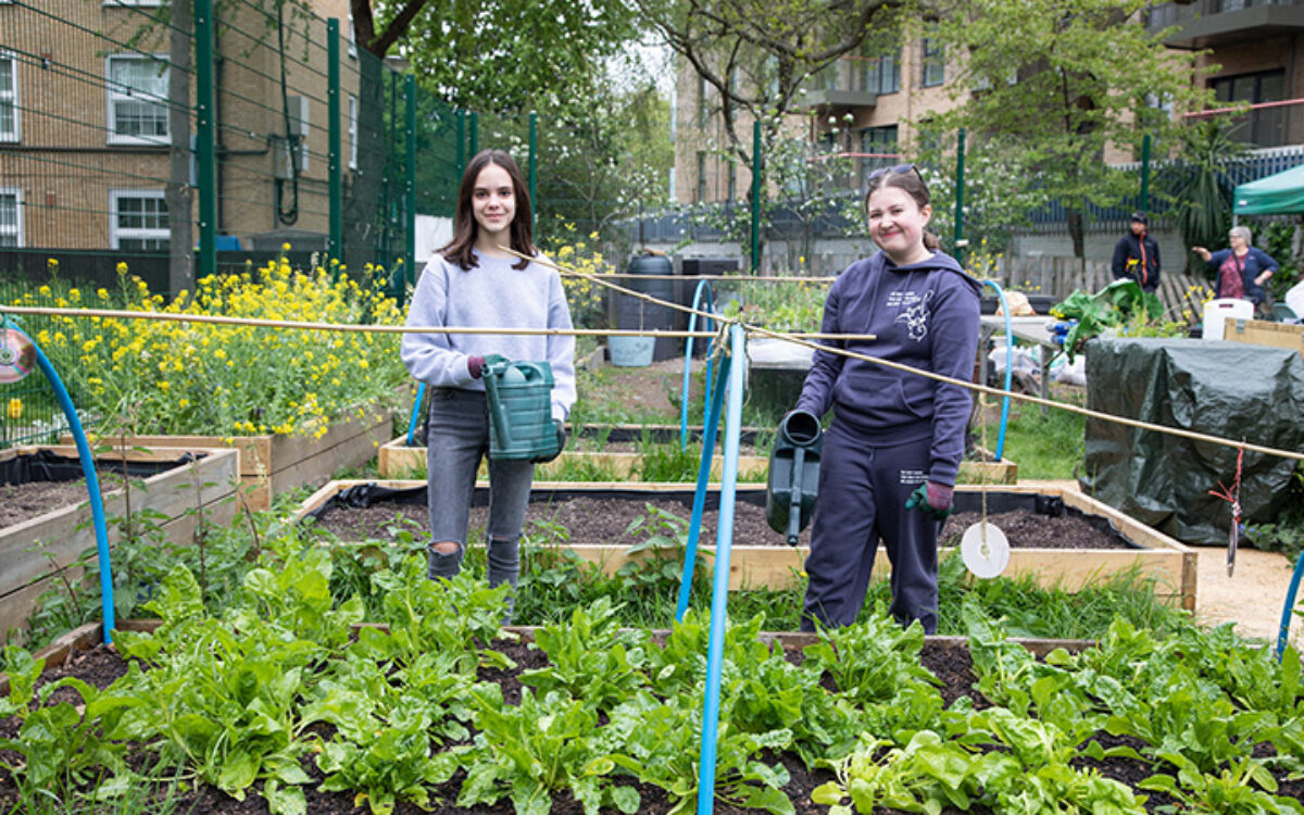 Two students with watering cans stood next to a bed of plants