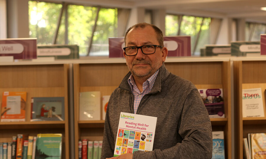 Tony Brown in a library holding a book called Reading Well For Mental Health