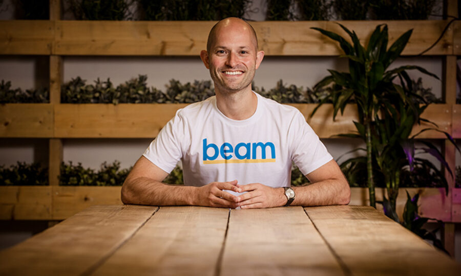 Alex Stephany, founder of Beam, sat at a table smiling wearing a t-shirt that says 'Beam'