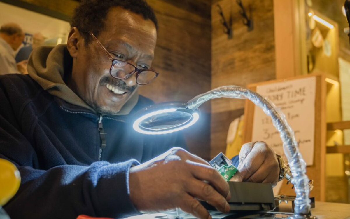 A Black man working to fix something under a bright lamp at a work station