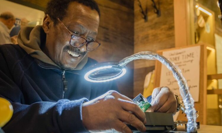 A Black man working to fix something under a bright lamp at a work station