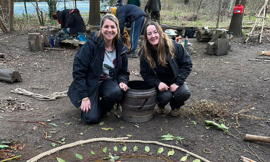 Childminders Danielle Franklin and Sarah Kenna crouched down in a wooded area with a pattern made from natural materials on the ground in front of them