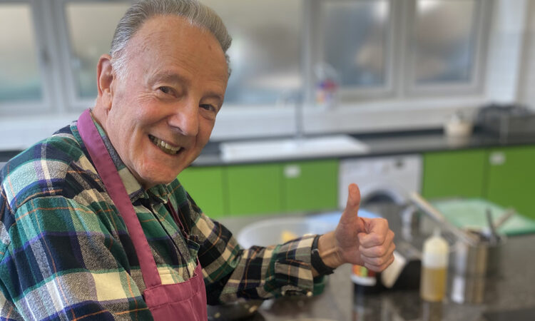 Older man wearing an apron in St Luke's Cookery School, smiling at the camera and doing a thumbs up