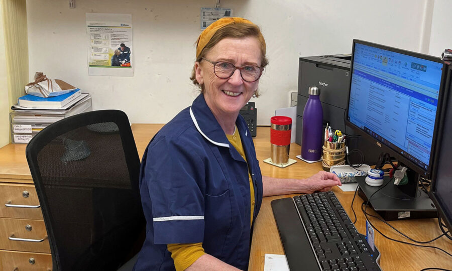 Breda Redmond-Hunt in her nurse's uniform sat smiling at a desk, in front of a computer