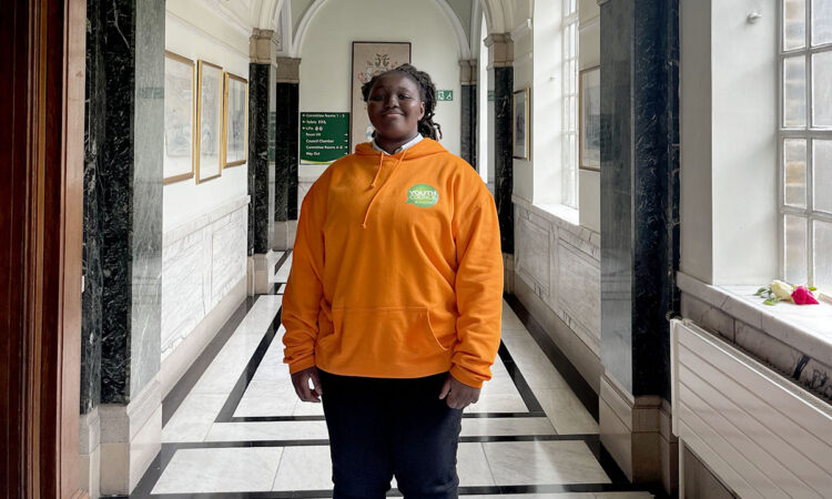 Youth councillor Maryam wearing an orange youth council hoody stood in the corridor of Islington Town Hall