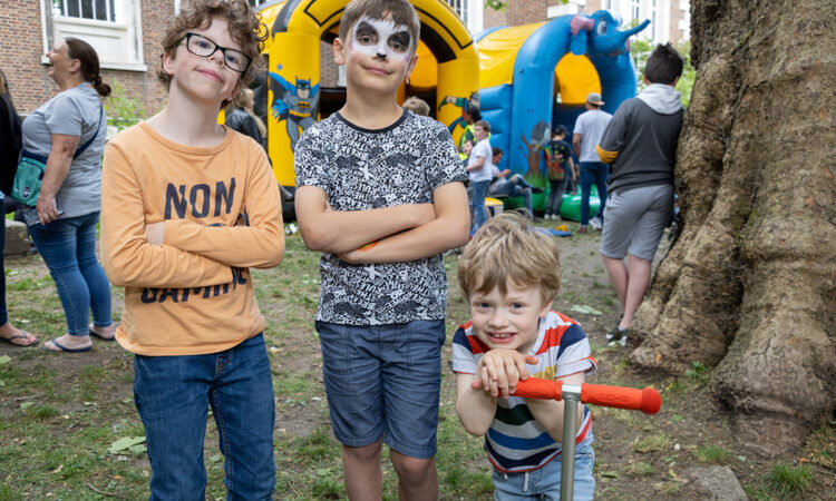 Three young boys, one on a scooter and one with facepaint on and a bouncy castle in the background