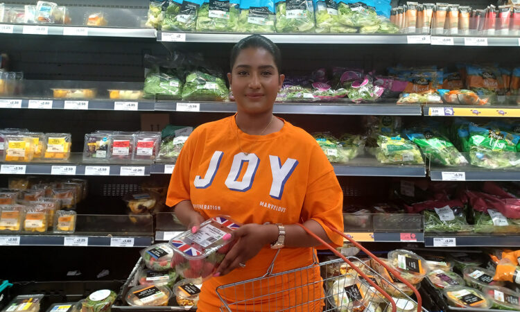 Fathima in a supermarket with salad and vegetables on the shelves behind her, holding a punnet of strawberries and shopping basket