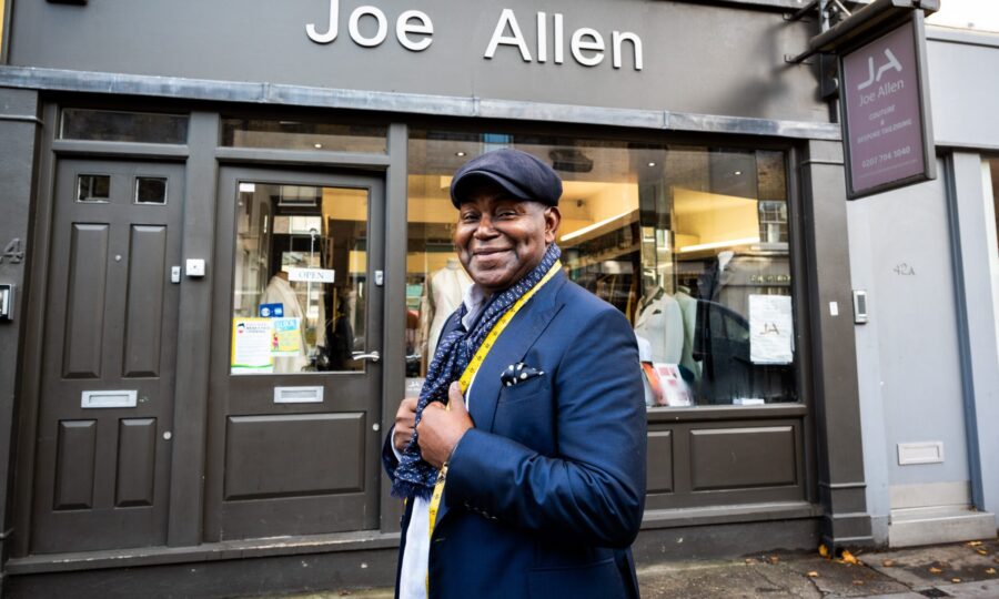 Joe Allen smiling at camera in front of his shop of the same name, wearing a suit and hat with a tape measure around his neck