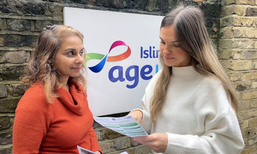Two women stood in front of a brick wall with an Islington Age UK sign, one looking and pointing at a piece of paper, the other facing her