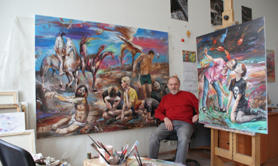 A man with a red top on is sat in a black chair in between two vibrant pieces of artwork, which hang from the wall and on a stand - he is looking towards the camera sat in a studio