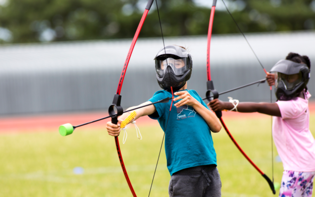 Two children with helmets on in a field doing archery