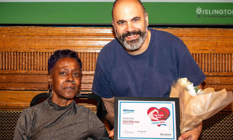 Shopay stood next to a Black woman in a wheelchair, holding his Dignity in Care award certificate in front of a stage in Islington Assembly Hall