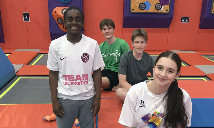 Four teenagers, a white girl, a black boy and two white boys, among the colourful trampolines at Sobell, smiling to camera