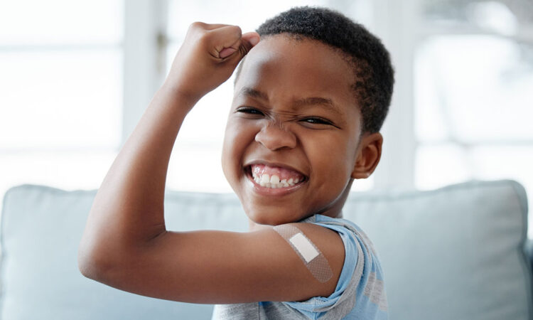 Young boy looking at the camera with a cheesy grin, holding his arm up in a 'strong' position with a plaster on his upper arm