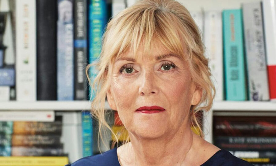 Head and shoulders shot of author Kate Atkinson, a middle-aged white woman with blonde curly hair, in front of shelves of books