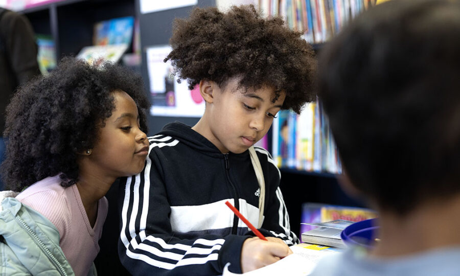 Two children, a boy and a girl, in a library looking down at the boy colouring