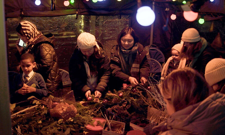 People gathered round a table with festoon lights above it making Christmas wreaths