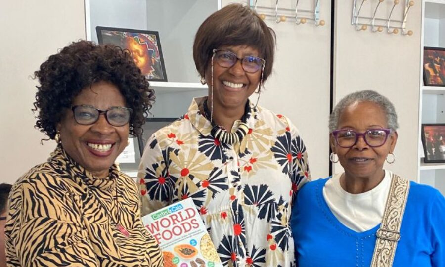 Three Black ladies smiling at the camera, all wearing glasses, the one in the middle holds a book called 'World Foods'