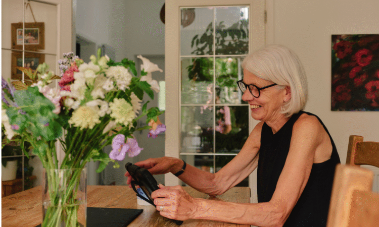 Older woman with grey hair smiling at an iPad while sitting on a wooden dining table with flowers on a vase next to her