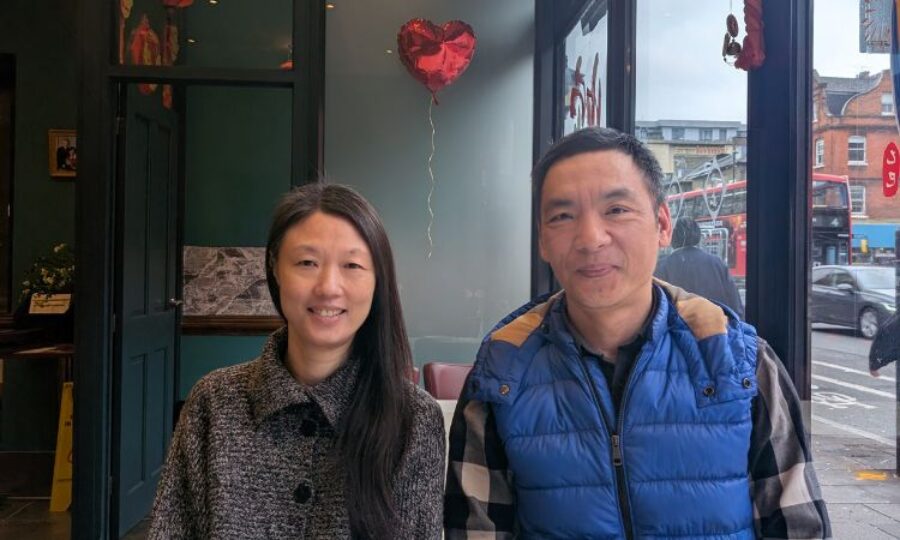 A lady with long dark hair wearing a grey buttoned up jacket is smiling at the camera sat next to a man with short dark hair wearing a blue jacket is also smiling at the camera with a love heart balloon in the background