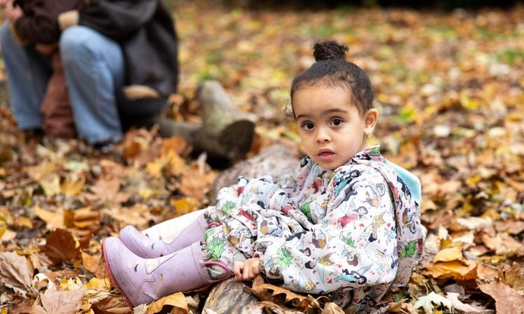 Young child in wellies sitting on the ground with autumn leaves