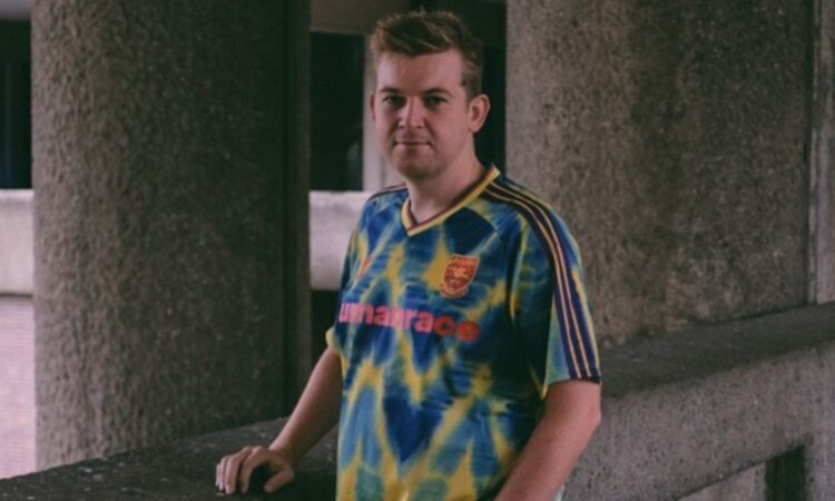 A man stands in an industrial looking setting looking at the camera while wearing a colourful Arsenal football top