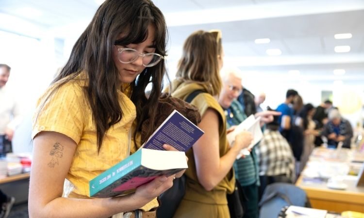 Woman with glasses wearing a yellow top in a library looking down at a book she's holding