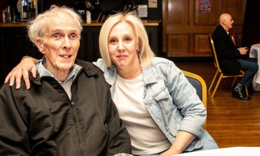 A woman and man sat smiling at the camera with the womans arm around the mans shoulder