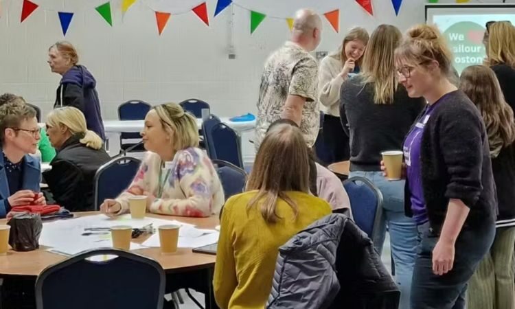 A group of people in a village hall, some sitting at round tables and some standing, chatting and having tea