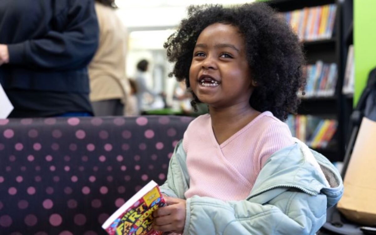 A little girl smiling excitedly at the camera while sitting in a library holding a book. She has dark curly hair