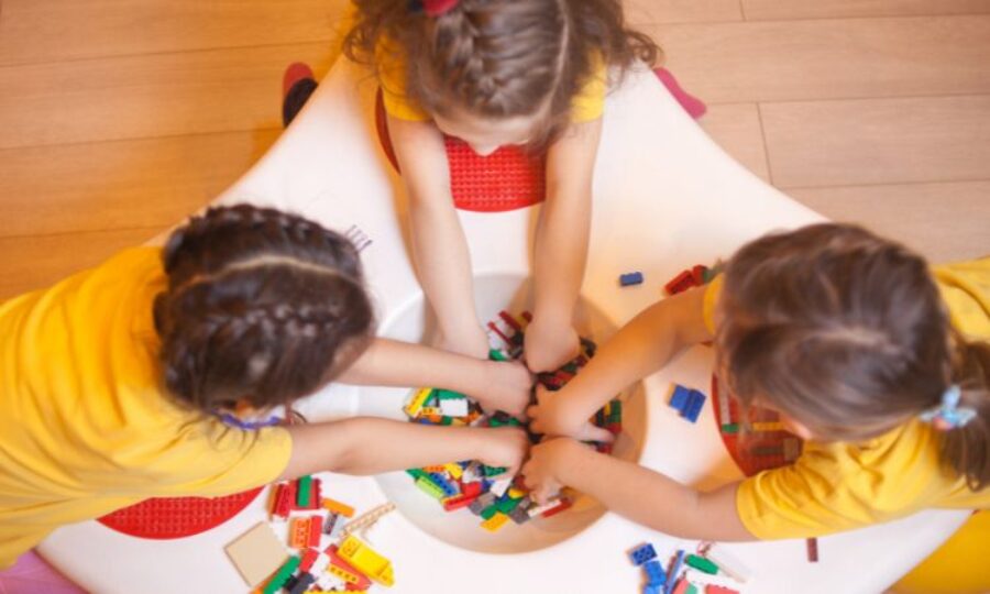three young girls playing with lego at a table. Angle is from above so you can only see their hair/ head not faces