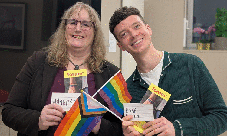 Two people smiling at the camera, one man and one woman, holding LGBTQ+ flags and Forum+ flyers