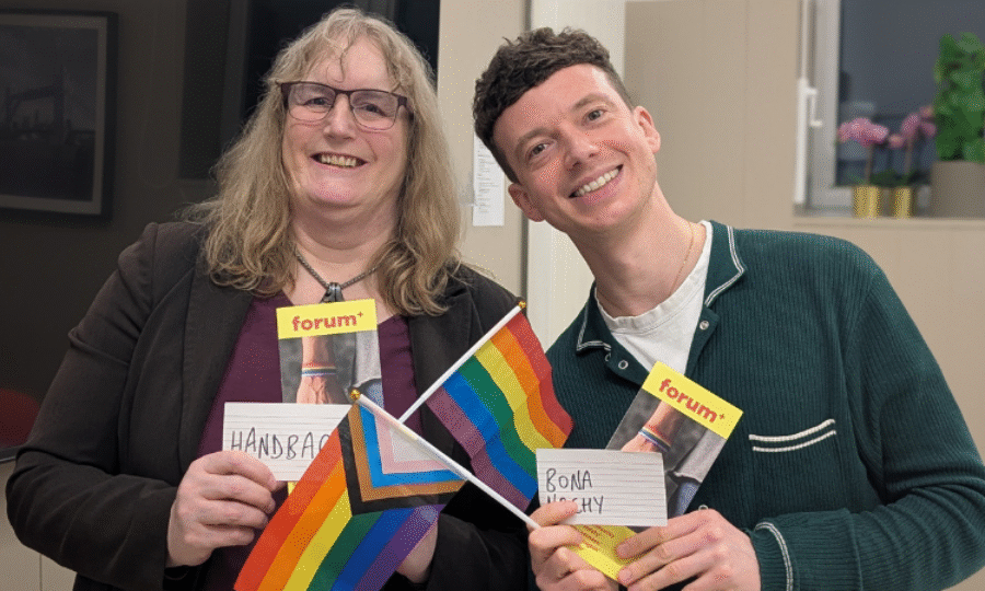Two people smiling at the camera, one man and one woman, holding LGBTQ+ flags and Forum+ flyers