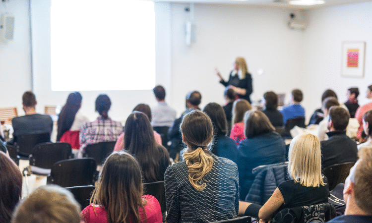 Speaker at the front of the room pointing to a screen while rows of people watch on