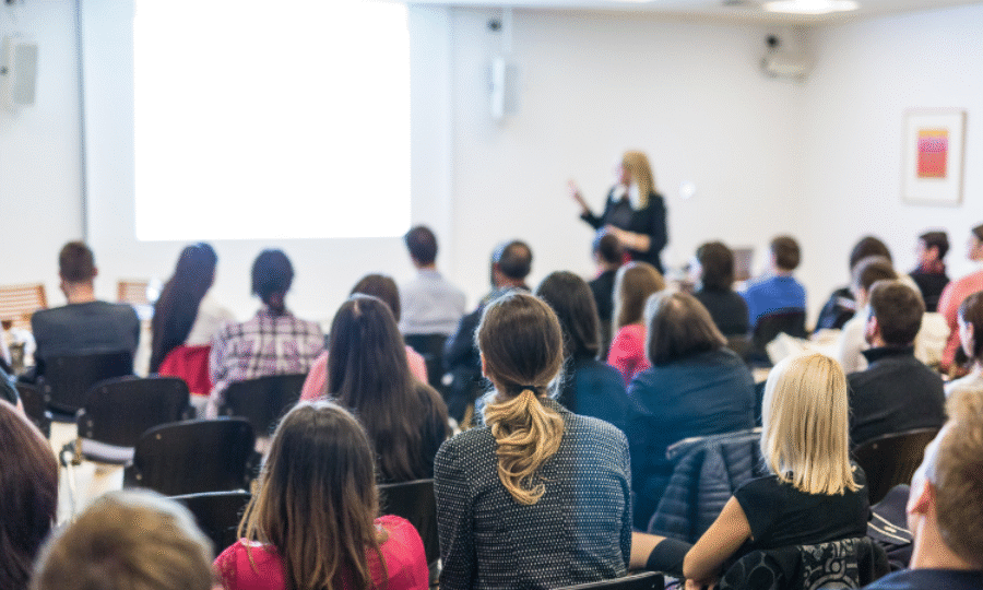 Speaker at the front of the room pointing to a screen while rows of people watch on