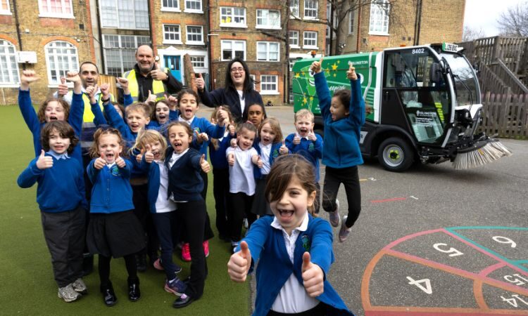 Lots of children standing in the playground in front of a brand new eco street sweeper. All the kids are smiling proudly and some of them have their thumbs up