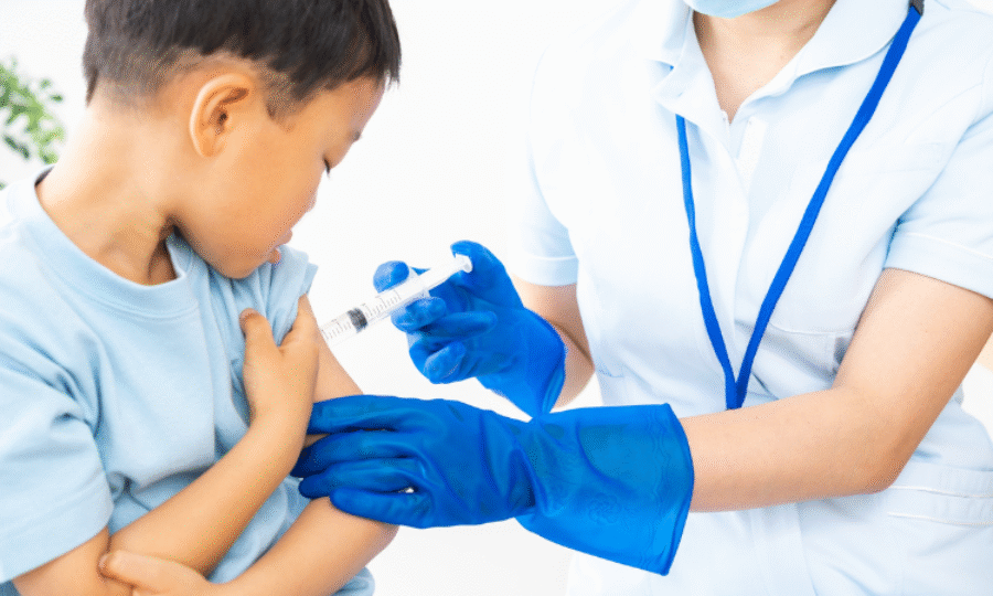 A young boy being vaccinated by a nurse who's face is not pictured just her upper body from the neck down