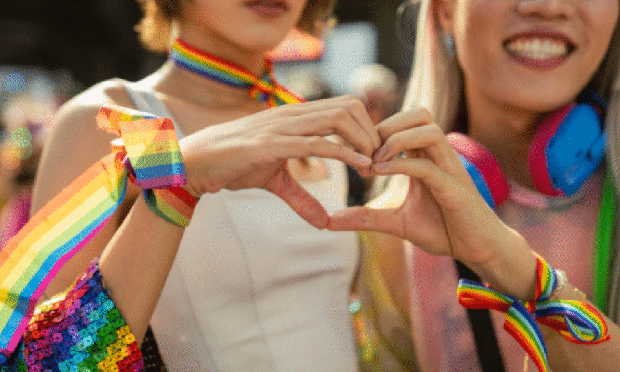 Two women dressed in LGBT flag and rainbow sequins doing a love heart with their hands