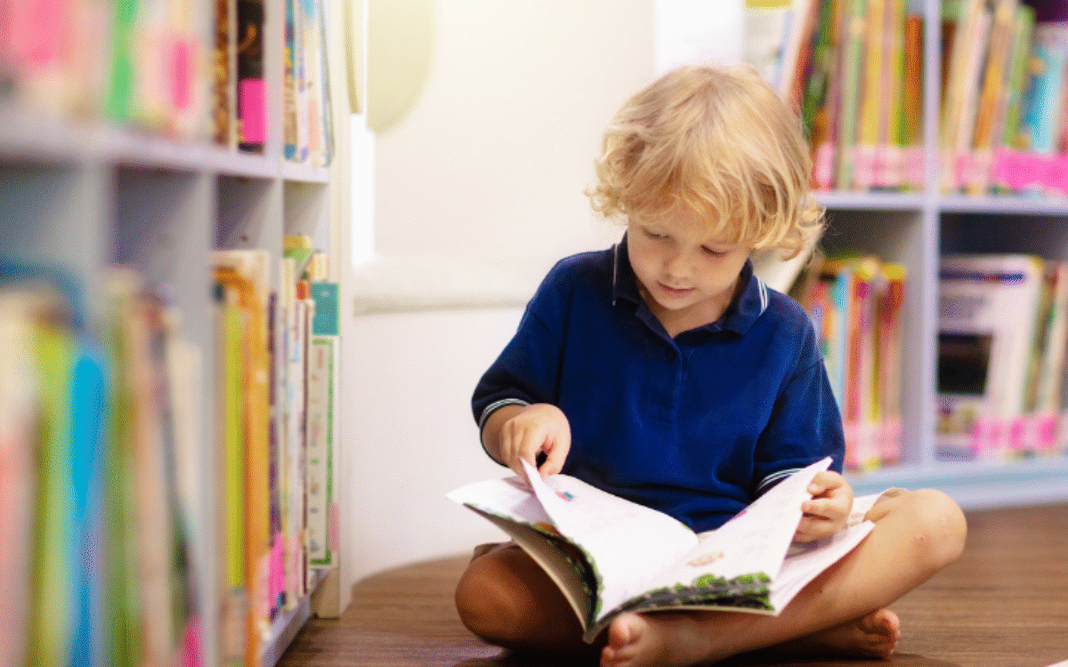 Young child with blonde hair sitting on the floor of a library reading a book with shelves of colourful books behind him