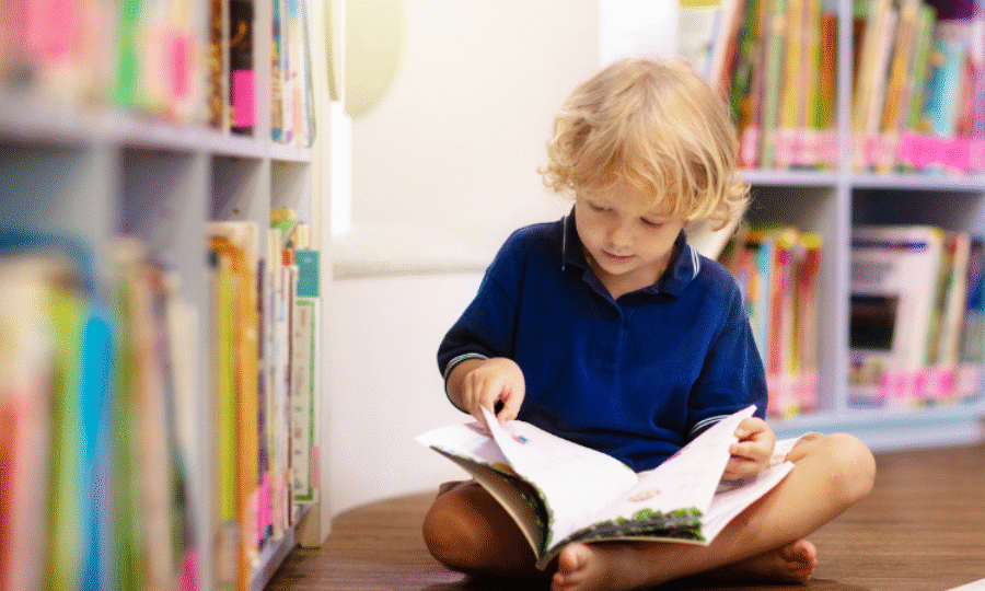 Young child with blonde hair sitting on the floor of a library reading a book with shelves of colourful books behind him